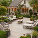 A beautifully rustic styled outdoor stone porcelain patio in front of a stone house, featuring a stone coffee table, black metal lounge chairs with cream cushions, a black dining table with chairs, and warm string lights throughout the garden.