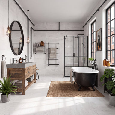The room has light white concrete-look tiled walls and floors, a woven jute rug, and large multi-pane windows. A modern industrial-style bathroom featuring a black clawfoot bathtub, a rustic wooden double-sink vanity with a gray countertop, a round black-framed mirror, and a black metal-framed glass shower enclosure. 