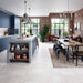 A modern open-plan kitchen and dining room interior. Large white concrete style porcelain floor tiles with a large concrete-look island, blue cabinetry, brick walls, and a dining table with black chairs on a beige rug.